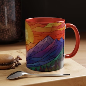 May include: Red ceramic mug with a stained glass design of a mountain range and a sunset. The mountains are purple, blue, and green, and the sunset is orange, yellow, and red.