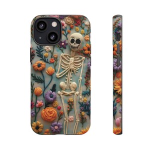 Puede incluir: Una funda de teléfono con un diseño de bordado floral colorido que presenta un esqueleto en el centro. El esqueleto está hecho de hilo blanco y está en una pose con las manos juntas. Las flores son de varios colores, incluyendo rosa, amarillo, naranja y morado.