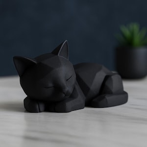 Modern black 3D printed low poly cat sculpture in a sleeping pose on a minimalist desk with art supplies and a bust in the background