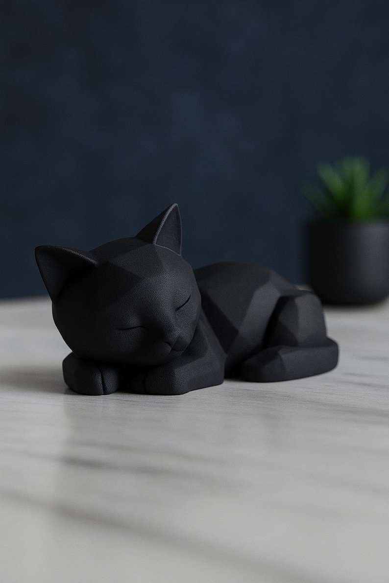 Modern black 3D printed low poly cat sculpture in a sleeping pose on a minimalist desk with art supplies and a bust in the background