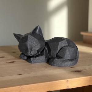 Modern black 3D printed low poly cat sculpture in a sleeping pose on a minimalist desk with art supplies and a bust in the background