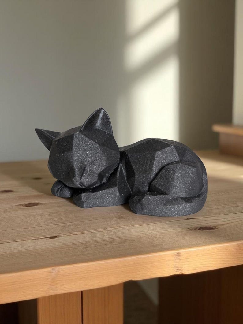 Modern black 3D printed low poly cat sculpture in a sleeping pose on a minimalist desk with art supplies and a bust in the background