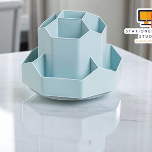 May include: A light blue desktop organizer with multiple compartments. The organizer has a hexagonal design and a rotating base. The text "STATIONERYSANC STUDIO DESKTOP ORGANIZER" is visible on the right side of the image.