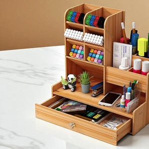 May include: A multi-compartment wooden desk organizer with a drawer. It contains various office supplies, including pens, markers, and small containers. The organizer also holds a small plant, a stapler, and a smartphone. The drawer has a crystal knob.