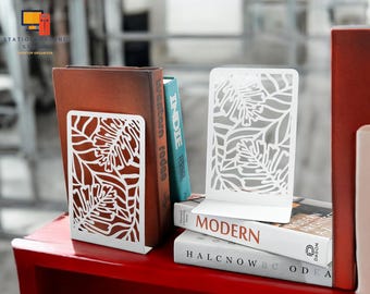 May include: Two white metal bookends with a leafy design. The bookends are holding up a stack of books with titles like "Indie" and "Modern".