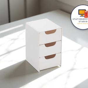 以下が含まれることがあります： ミニマリストデザインの白い3段引き出しデスクトップオーガナイザー。各引き出しには、湾曲したライトブラウンのハンドルが付いています。オーガナイザーは、しっかりとした不透明な素材でできており、白い表面に置かれています。 StationerySanc Studioのロゴが背景にあります。