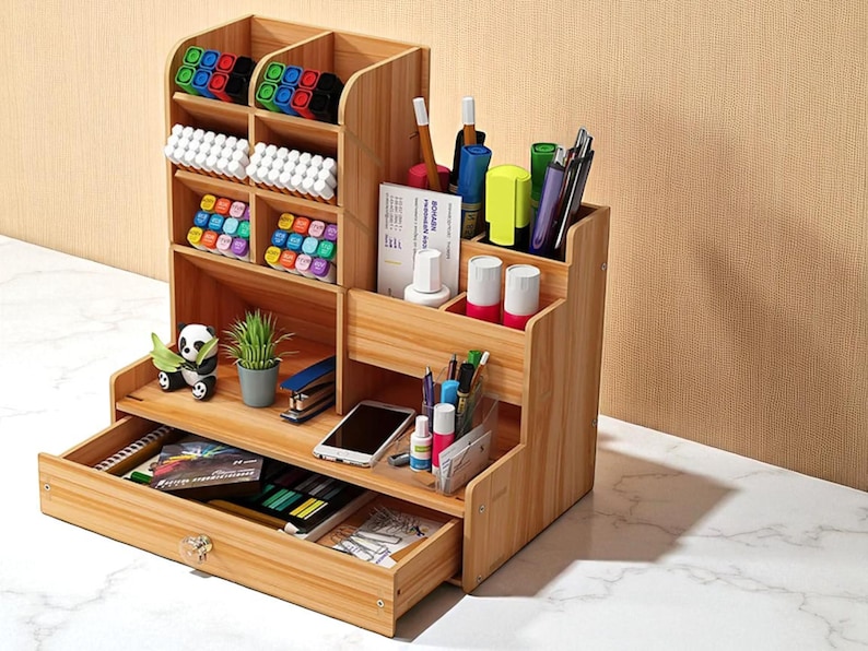 May include: A multi-tiered wooden desk organizer with multiple compartments for pens, markers, and other office supplies. The organizer has a drawer and is filled with various items, including a small panda figurine and a potted plant.