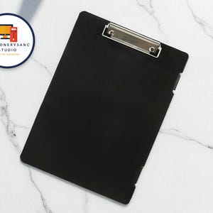 May include: A black clipboard with a silver metal clip, on a white marble surface. The clipboard is rectangular and has a smooth finish. A circular logo with a computer and text is in the upper left corner.