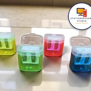 Könnte beinhalten: Vier farbige Anspitzer in Gelb, Grün, Rot und Blau. Jeder Anspitzer hat einen transparenten Kunststoffkörper mit farblich passendem Innenraum und einem Klappdeckel. Die Anspitzer sind auf einer hellen Oberfläche angeordnet. Das Bild enthält ein Logo mit der Aufschrift "STATIONERYSANC STUDIO DESKTOP ORGANIZER."