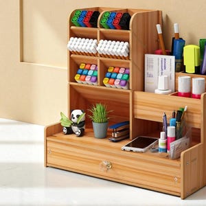 May include: A wooden desk organizer with multiple compartments and a drawer. It holds various office supplies, including pens, markers, and a small potted plant. A panda figurine sits on the bottom shelf. The organizer is made of light-colored wood.