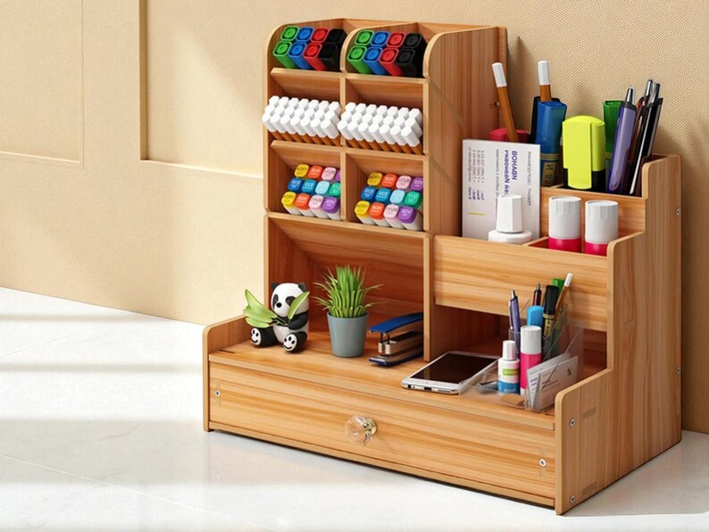 May include: A wooden desk organizer with multiple compartments and a drawer. It holds various office supplies, including pens, markers, and a small potted plant. A panda figurine sits on the bottom shelf. The organizer is made of light-colored wood.