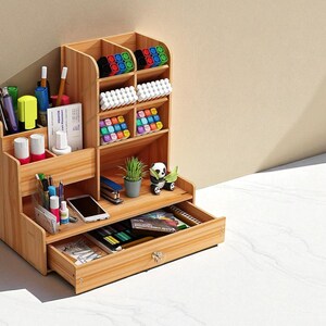 May include: A wooden desk organizer with multiple compartments and a drawer. It contains pens, markers, and other office supplies. The organizer has a natural wood finish and is designed to hold various stationery items, a phone, and small decorative items.
