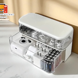 Puede incluir: Organizador de escritorio blanco y transparente con múltiples compartimentos. La sección superior contiene cables, mientras que la sección inferior contiene cargadores, pilas y otros accesorios. El organizador tiene el texto "STATIONERYSANC STUDIO DESKTOP ORGANIZER".