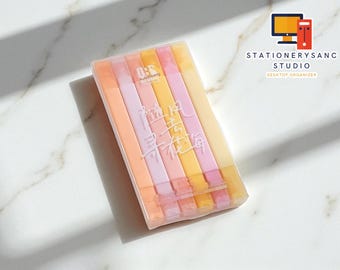 Può includere: Una custodia di plastica trasparente contenente sei evidenziatori dai colori pastello. Gli evidenziatori sono nelle tonalità del rosa, dell'arancione e del giallo. La custodia presenta caratteri cinesi stampati. L'immagine è su una superficie di marmo bianco.