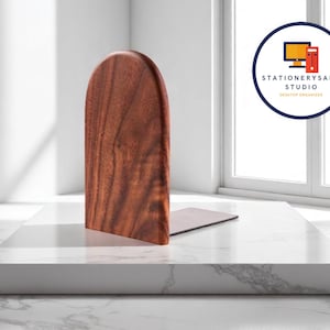 May include: A wooden bookend with a curved top, showcasing a rich brown wood grain. The bookend is positioned on a white marble surface, with a logo in the background that reads "STATIONERYSANC STUDIO DESKTOP ORGANIZER".