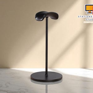 Può includere: Un supporto nero per cuffie con base rotonda e parte superiore curva. Il supporto è in metallo e ha un design minimalista. Il logo STATIONERYSANC STUDIO è visibile sullo sfondo.