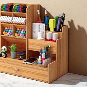 May include: A wooden desk organizer with multiple compartments and a drawer. It holds pens, markers, and other office supplies. A small panda figurine and a potted plant are also visible.