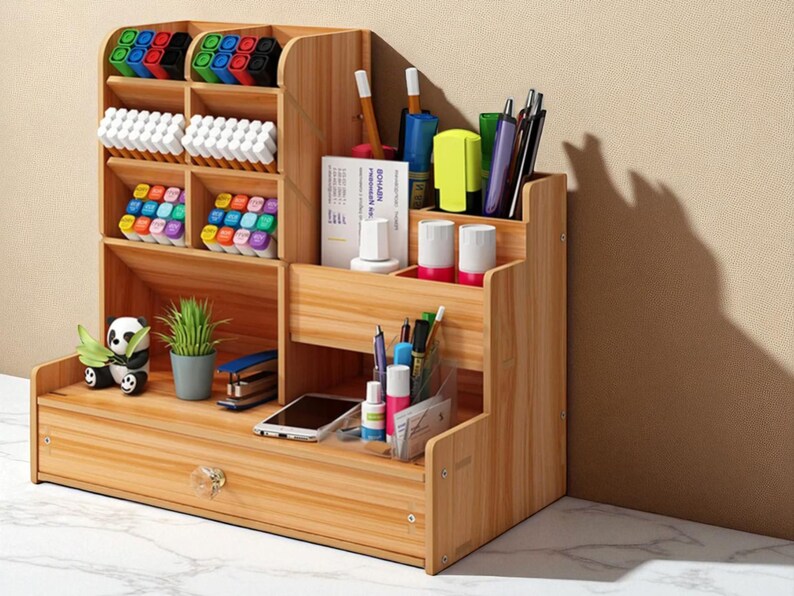 May include: A wooden desk organizer with multiple compartments and a drawer. It holds pens, markers, and other office supplies. A small panda figurine and a potted plant are also visible.