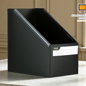 May include: A black desktop organiser with a slanted opening and a white label. The edges are trimmed with a silver metal. The text "STATIONERYSANC STUDIO DESKTOP ORGANIZER" is in the upper right corner.