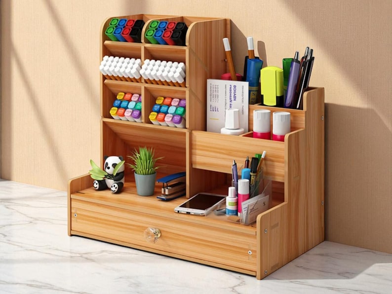 May include: Wooden desk organizer with multiple compartments for pens, markers, and other office supplies. Features a drawer, a small panda figurine, and a potted plant. The organizer is made of light-colored wood.