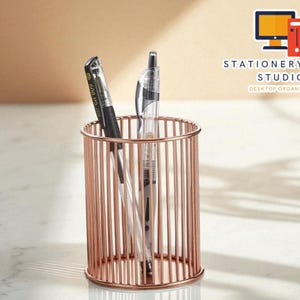 Puede incluir: Un portalápices de metal color oro rosa con barras verticales, que contiene dos bolígrafos. Un bolígrafo es negro y el otro transparente con punta plateada. El fondo es de color beige claro. El texto "STATIONERYSANC STUDIO DESKTOP ORGANIZER" es visible.