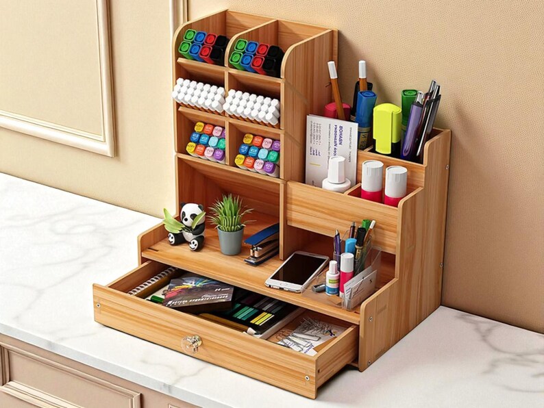 May include: A wooden desk organizer with multiple compartments and a drawer. It contains various office supplies, including pens, markers, and small plants. The organizer is made of light-colored wood and sits on a white surface.