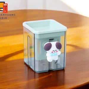 Peut inclure: Un organisateur de bureau en plastique bleu clair avec un devant transparent et un motif de chien de dessin animé mignon. L'organisateur est étiqueté "Stationery Sanc Studio Desktop Organizer".