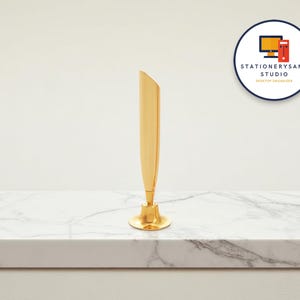 Op de afbeelding: Een goudkleurige pennenhouder met een uitlopende basis en een strakke, schuine bovenkant. De pennenhouder staat op een wit marmeren oppervlak. Het logo "STATIONERYSANC STUDIO DESKTOP ORGANIZER" is zichtbaar.