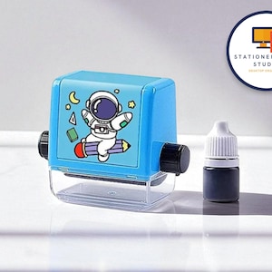 May include: A blue self-inking stamp featuring an astronaut cartoon. The stamp has black knobs and a clear base. A small bottle of black ink is next to the stamp. The image also includes the text "STATIONERYSANC STUDIO DESKTOP ORGANIZER".