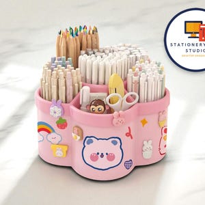 Op de afbeelding: Roze bureau-organizer met meerdere compartimenten gevuld met pennen, potloden en andere kantoorbenodigdheden. De organizer is versierd met schattige cartoonfiguren en een regenboog. De tekst "STATIONERYSANC STUDIO DESKTOP ORGANIZER" staat in een cirkel.