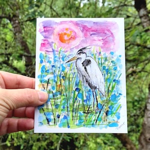 May include: A watercolor painting on a rectangular card featuring a grey heron standing among blue and green reeds. The background displays a vibrant pink and purple sky with a bright orange sun. The artist signature Tamar is in the corner.