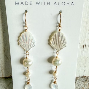 May include: A pair of dangling earrings with a seashell design, featuring iridescent white shells, freshwater pearls, and a teardrop-shaped stone. The earrings are suspended from gold-colored hooks. The card behind the earrings says "Hana Lima Maui MADE WITH ALOHA."