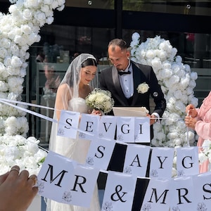Könnte beinhalten: Ein Brautpaar steht vor einem weißen Blumenbogen und hält ein Banner mit der Aufschrift "SEVGI SAYG MR & MRS". Die Braut trägt ein weißes Kleid und einen Schleier, der Bräutigam einen schwarzen Smoking. Eine Hochzeitszeremonie im Freien.