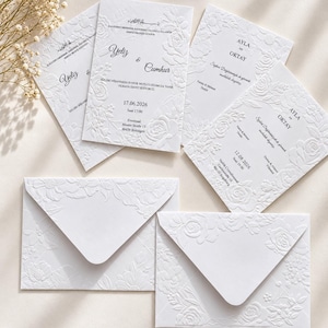 Könnte beinhalten: Eine Sammlung weißer Hochzeitseinladungskarten und passender Umschläge mit geprägten Blumenmustern. Die Karten zeigen gedruckten Text, darunter Namen wie Yeliz, Cumhur, Ayla und Oktay, sowie Daten, Uhrzeiten und Veranstaltungsorte.