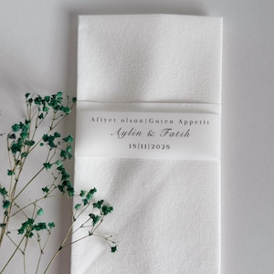 May include: A white linen napkin, folded and secured with a translucent band. The band displays the text "Afiyet olsun|Guten Appetit", names "Aylin & Fatih", and the date "15|11|2025". Green floral elements are also present.