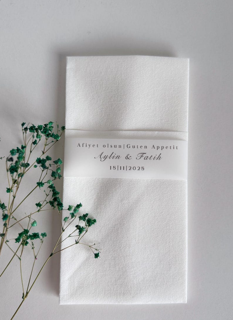 May include: A white linen napkin, folded and secured with a translucent band. The band displays the text "Afiyet olsun|Guten Appetit", names "Aylin & Fatih", and the date "15|11|2025". Green floral elements are also present.