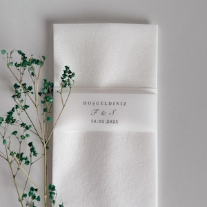 May include: A white rectangular invitation card with a translucent band featuring the text "HOŞGELDİNİZ", initials "F & S", and the date "10.05.2025". A sprig of green foliage is on the left side of the card.