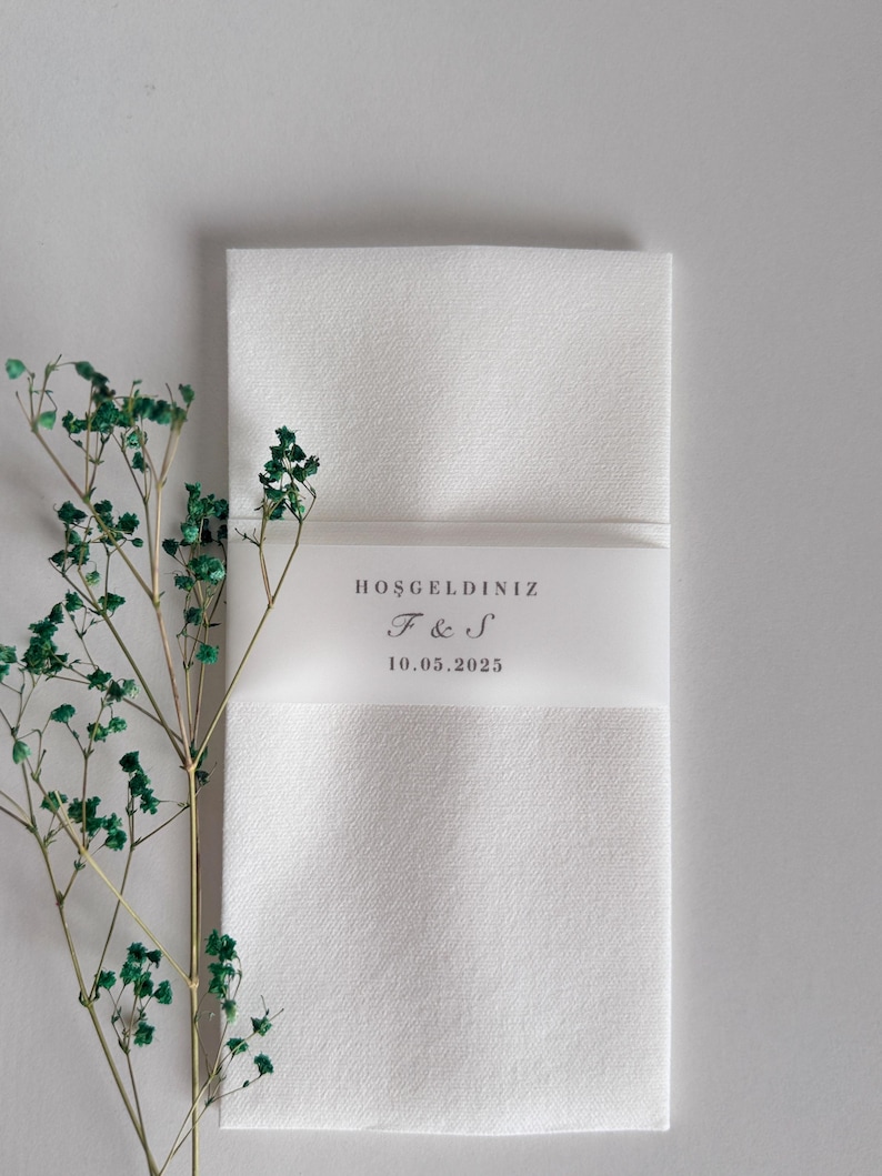 May include: A white rectangular invitation card with a translucent band featuring the text "HOŞGELDİNİZ", initials "F & S", and the date "10.05.2025". A sprig of green foliage is on the left side of the card.