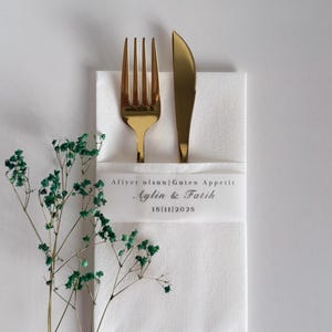 May include: A gold fork and knife sit on a white napkin, with a sprig of green foliage to the left. The napkin has a white band with the text "Afiyet olsun|Guten Appetit Aylin & Fatih 15|11|2025".