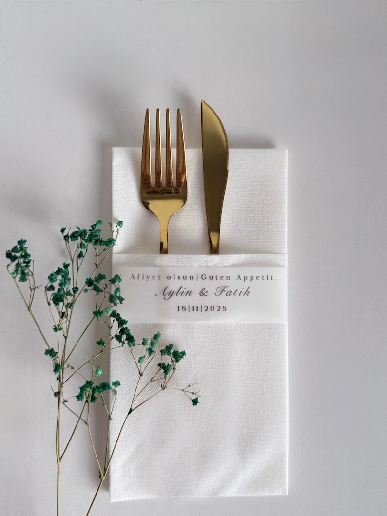 May include: A gold fork and knife sit on a white napkin, with a sprig of green foliage to the left. The napkin has a white band with the text "Afiyet olsun|Guten Appetit Aylin & Fatih 15|11|2025".