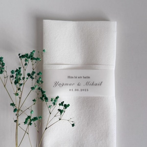 May include: A folded white linen napkin with a decorative band featuring the text "H&ucirc;n bi x&ecirc;r hatin, Yağmur & Mikail, 01.06.2025". Green sprigs of dried flowers are placed to the left of the napkin.
