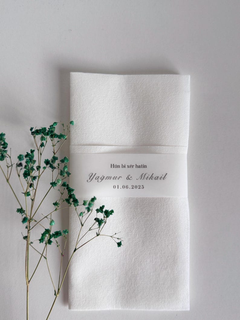 May include: A folded white linen napkin with a decorative band featuring the text "H&ucirc;n bi x&ecirc;r hatin, Yağmur & Mikail, 01.06.2025". Green sprigs of dried flowers are placed to the left of the napkin.