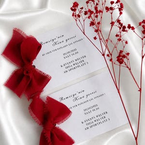 May include: Two red fabric bows with frayed edges and two white cards with black text. The cards have event details, including a date and time. Red dried flowers are arranged on the right side of the image. The background is a white, silky fabric.