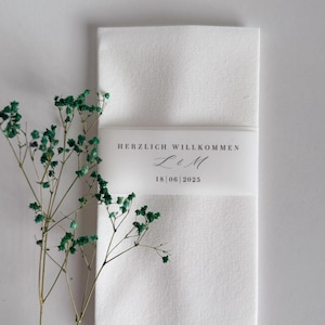 May include: A folded white linen napkin with a translucent band bearing the text "HERZLICH WILLKOMMEN" and the date "18 | 06 | 2025". Green dried flowers are positioned to the left of the napkin. A wedding or event detail.