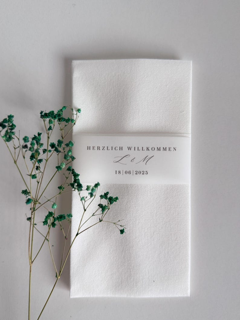 May include: A folded white linen napkin with a translucent band bearing the text "HERZLICH WILLKOMMEN" and the date "18 | 06 | 2025". Green dried flowers are positioned to the left of the napkin. A wedding or event detail.