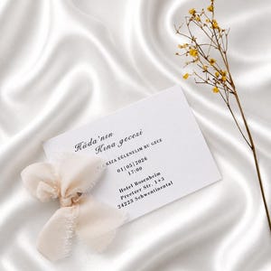 May include: A white invitation card with text and a cream-colored ribbon bow, set against a white satin background. The card includes event details and is accompanied by a sprig of yellow flowers.