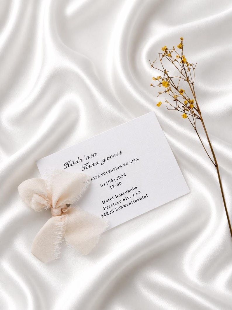 May include: A white invitation card with text and a cream-colored ribbon bow, set against a white satin background. The card includes event details and is accompanied by a sprig of yellow flowers.
