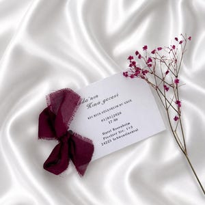 May include: A white card with burgundy ribbon and text, resting on a white satin surface. The card includes event details: date, time, and location. A sprig of small, purple flowers is placed near the card.
