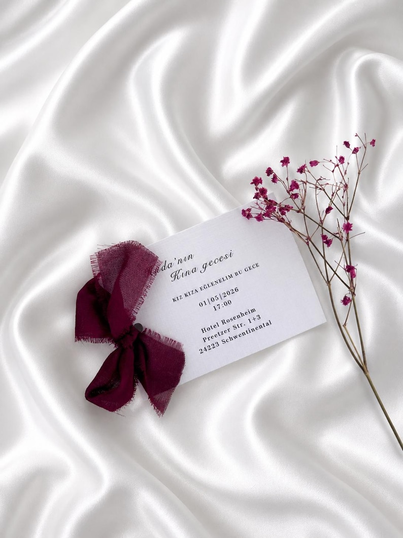 May include: A white card with burgundy ribbon and text, resting on a white satin surface. The card includes event details: date, time, and location. A sprig of small, purple flowers is placed near the card.