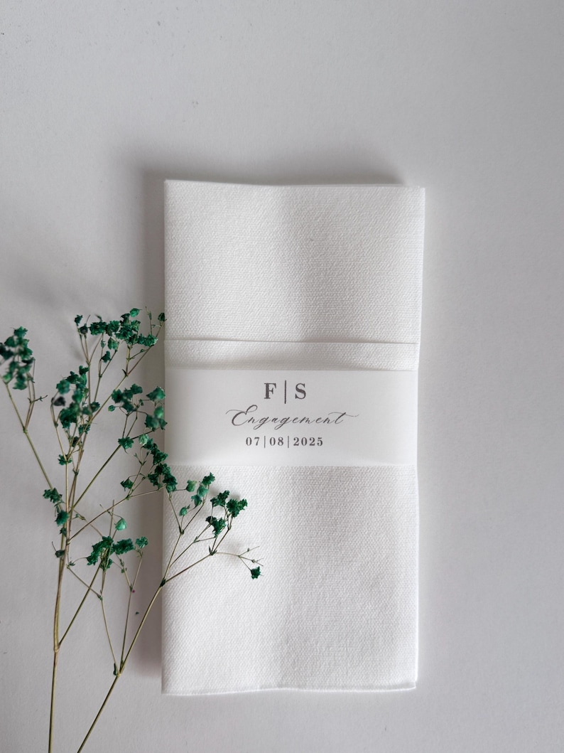 May include: A folded white linen napkin with a decorative band. The band features the initials "F|S", the word "Engagement", and the date "07|08|2025". Green sprigs of dried flowers are to the left of the napkin. The napkin is likely for a wedding or engagement.